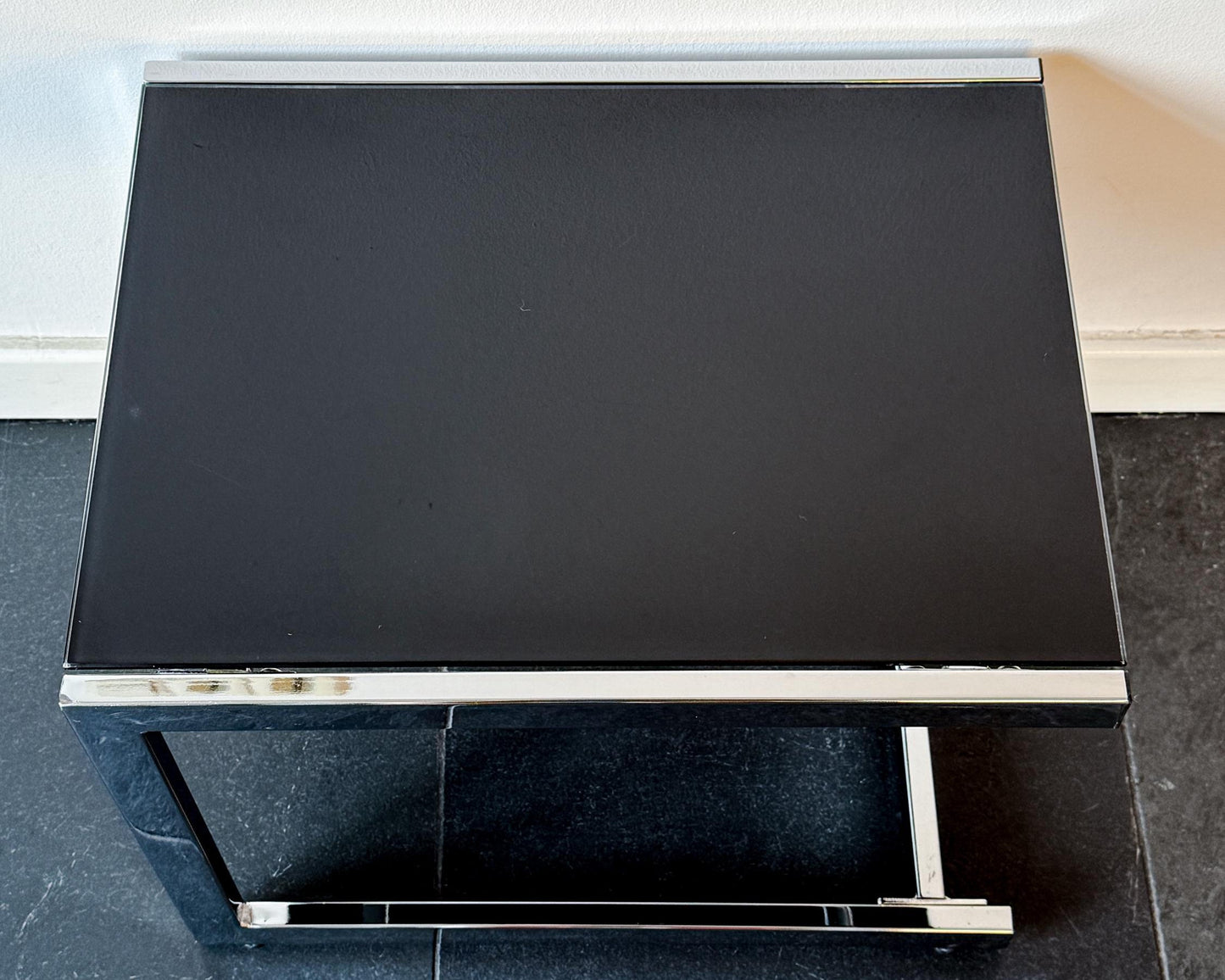 Black glass coffee table with chrome frame on a dark tiled floor.