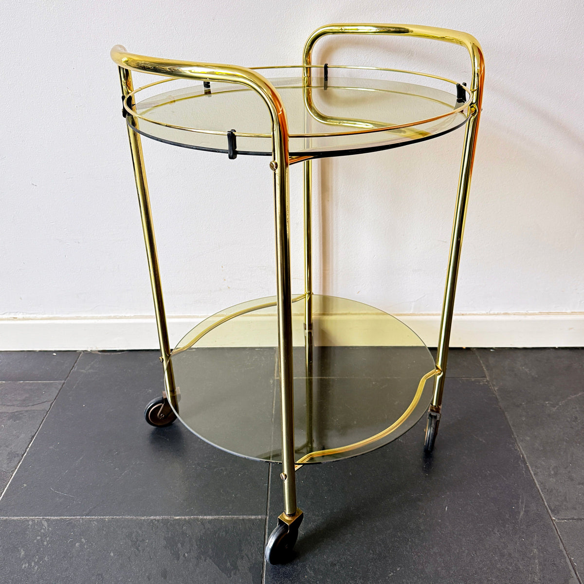 Mid-century modern brass bar cart with two circular smoked glass shelves, curved brass handles and four castor wheels.